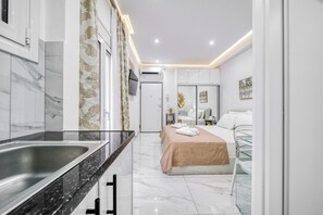 Une chambre d’hôtel moderne dotée d’une salle de bain, d’un lit recouvert d’une couverture beige, d’une cabine de douche en verre et d’un plafond blanc avec des spots encastrés.