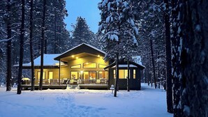 Un chalet doté d’une grande terrasse et d’un bâtiment séparé, le tout entouré d’arbres couverts de neige.