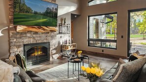 Un salon moderne avec une cheminée en pierre, un grand téléviseur à écran plat et une vue sur un terrain de golf par les fenêtres.