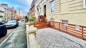 Una calle estrecha con coches aparcados y un edificio con valla de madera y zona de grava.