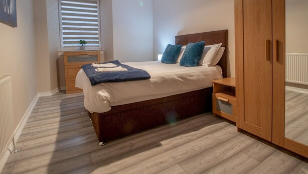 Un dormitorio con cama, mesitas de noche, armario, ventana con persianas e iluminación en el techo.