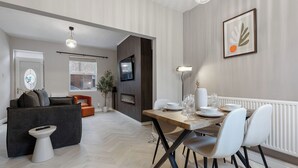 A modern living room with a sofa, a dining table set for four, and a TV mounted on the wall. - 29 Carleton Street · Newly Refurbished 2-Bed Terraced Home in York - Pass the Keys (York)