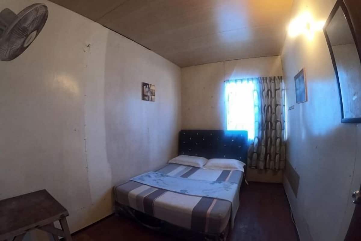 Habitació petita amb un llit individual, un panell frontal de fusta, una tauleta de nit, un ventilador fixat a la paret i una finestra amb cortines.