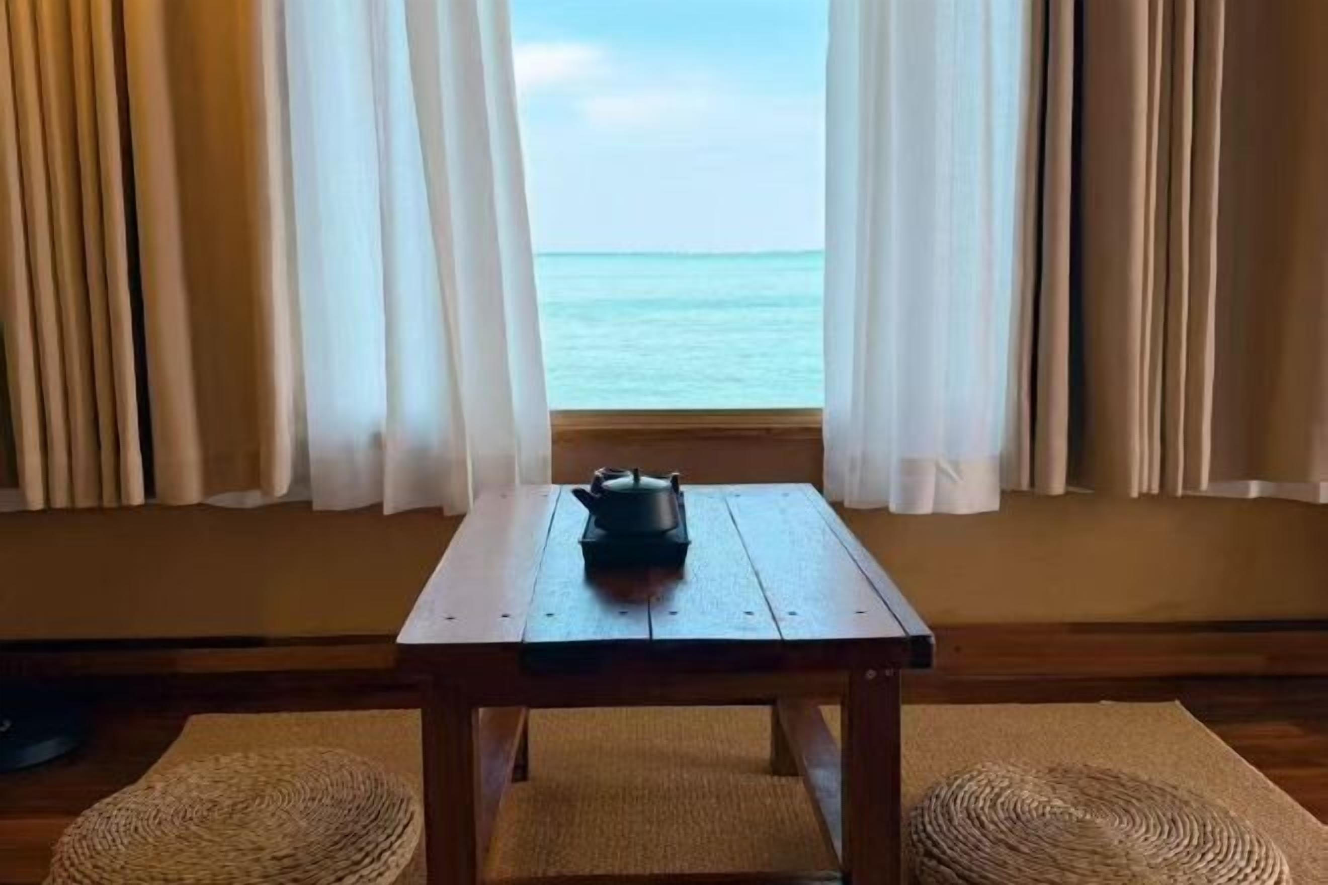 Een kamer met een houten tafel, een geweven mat en uitzicht op de oceaan door dunne gordijnen.