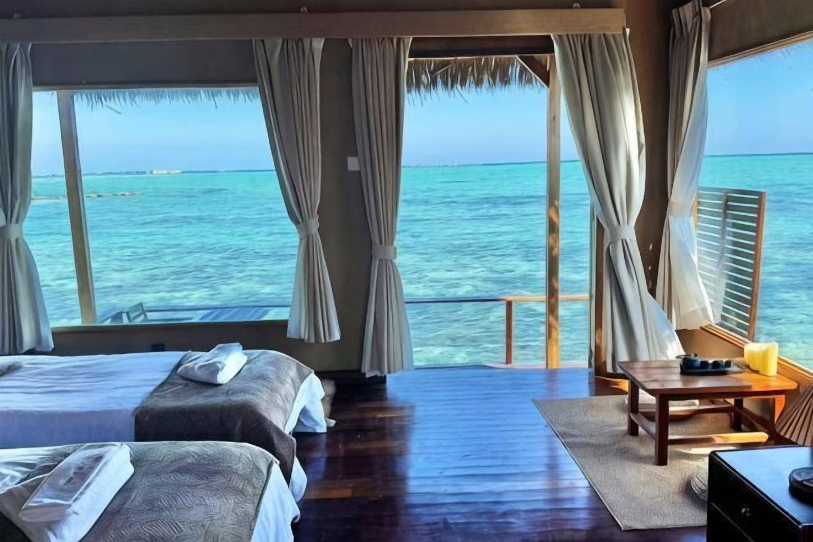 Una habitación con dos camas, piso de madera, una mesa con una silla y vista al océano.