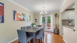 Comedor con mesa de madera oscura y sillas grises, una lámpara de araña y vistas a la cocina y zona exterior.