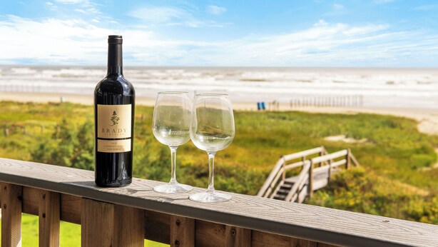 Una botella de vino y dos copas sobre una terraza de madera con vista a una playa.