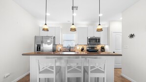 Una cocina moderna con armarios blancos, una isla central con taburetes de bar, electrodomésticos de acero inoxidable e iluminación colgante.