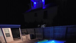 Ein rechteckiger Swimmingpool mit blauer Beleuchtung, umgeben von einem Holzzaun und einer Pflasterterrasse.