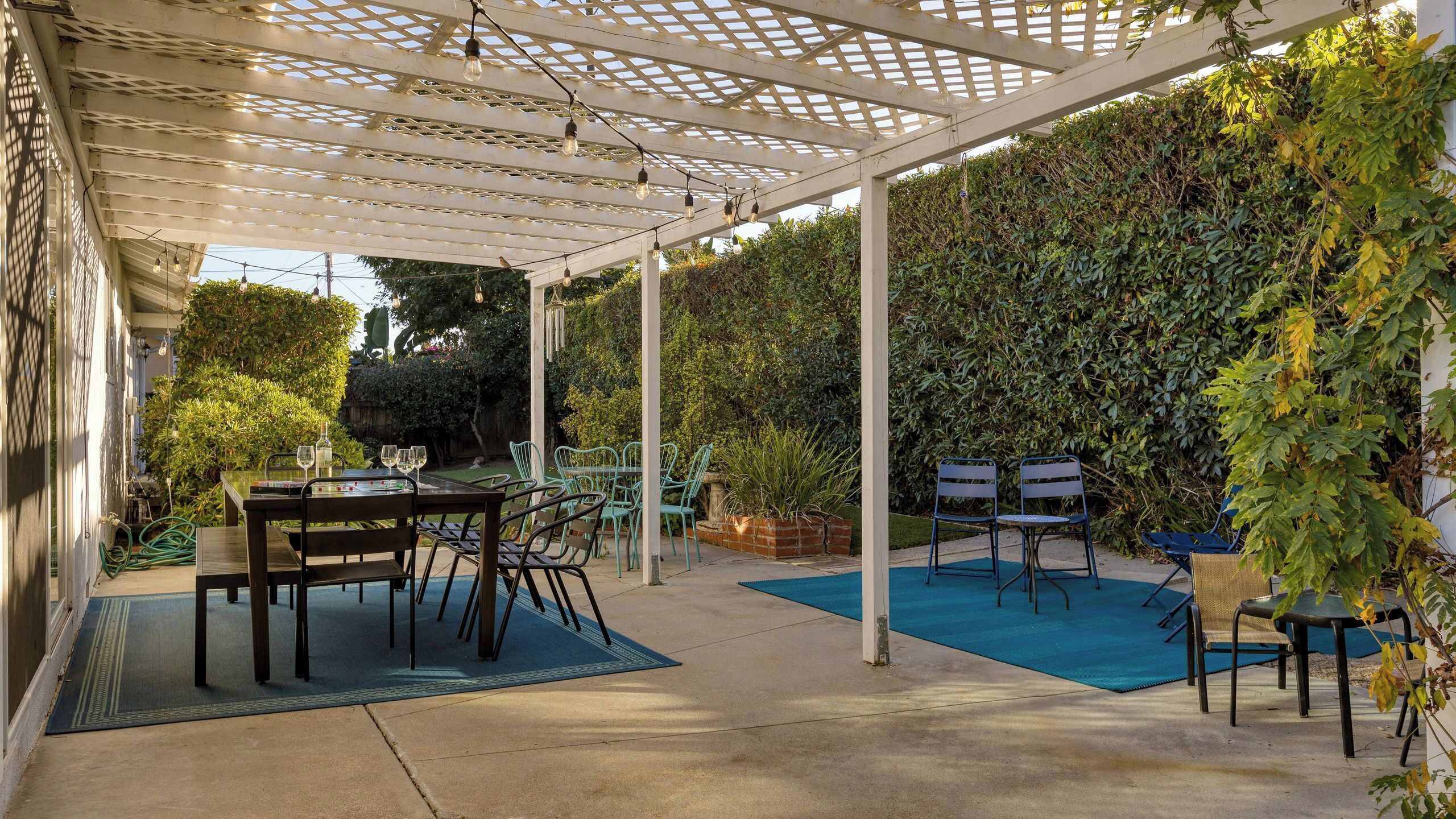 A covered patio with tables and chairs, a pool, and a lush green hedge.