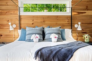 Een net opgemaakt bed met een deken in witte en blauwe strepen, twee witte handdoeken met roze bloemen, en een vaas met zonnebloemen op een nachtkastje.