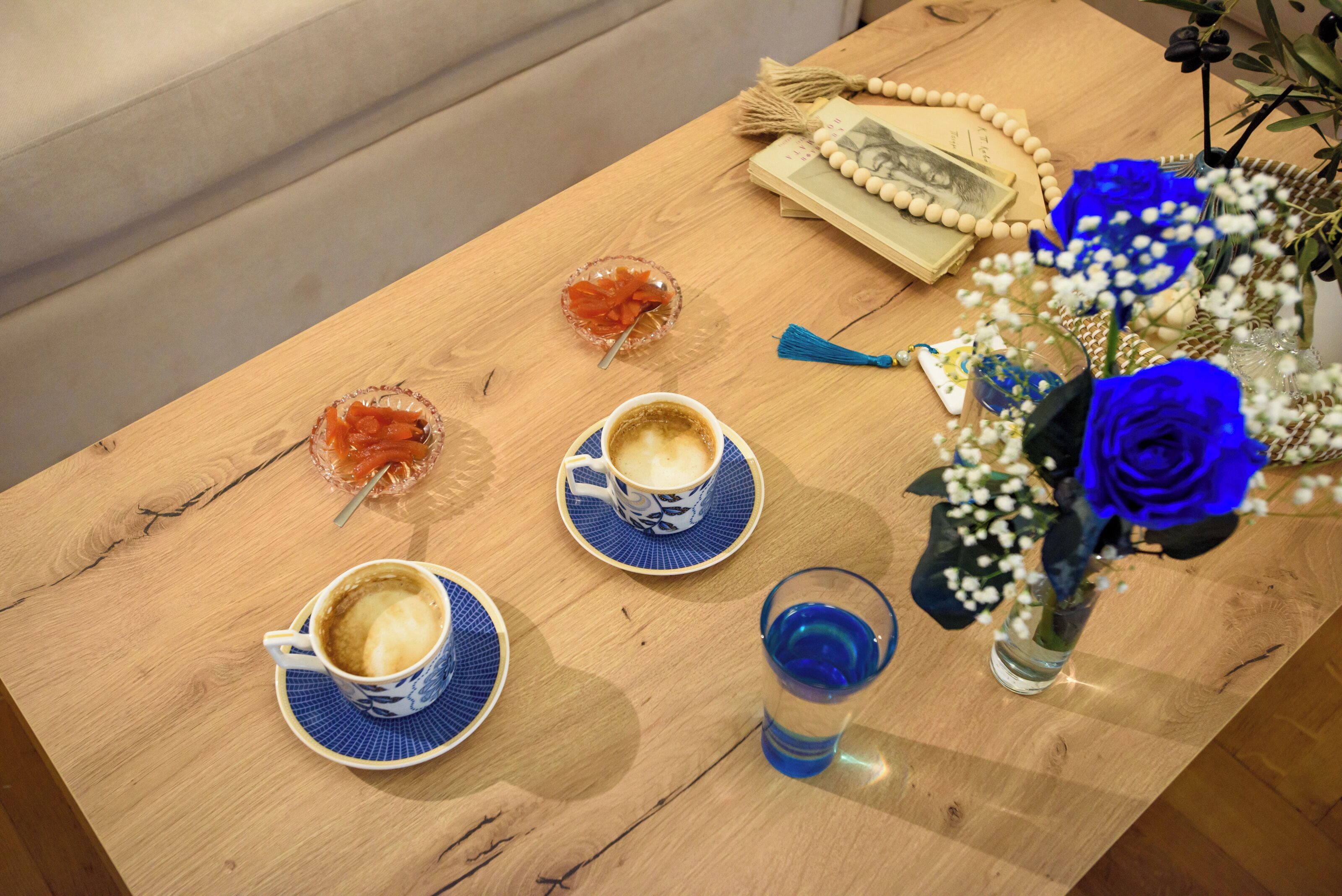 A wooden table with two cups of coffee, a glass of blue liquid, a bowl of orange fruit, and a vase with blue roses.