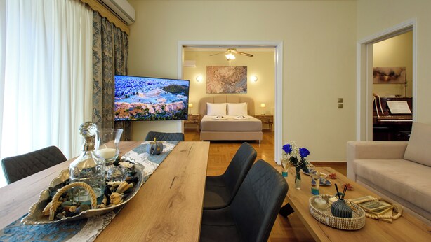 A modern living room with a wooden dining table, black leather chairs, a flat-screen TV, a sofa, and a piano. - Blue Ark Acropolis Signature Residence (Athens)