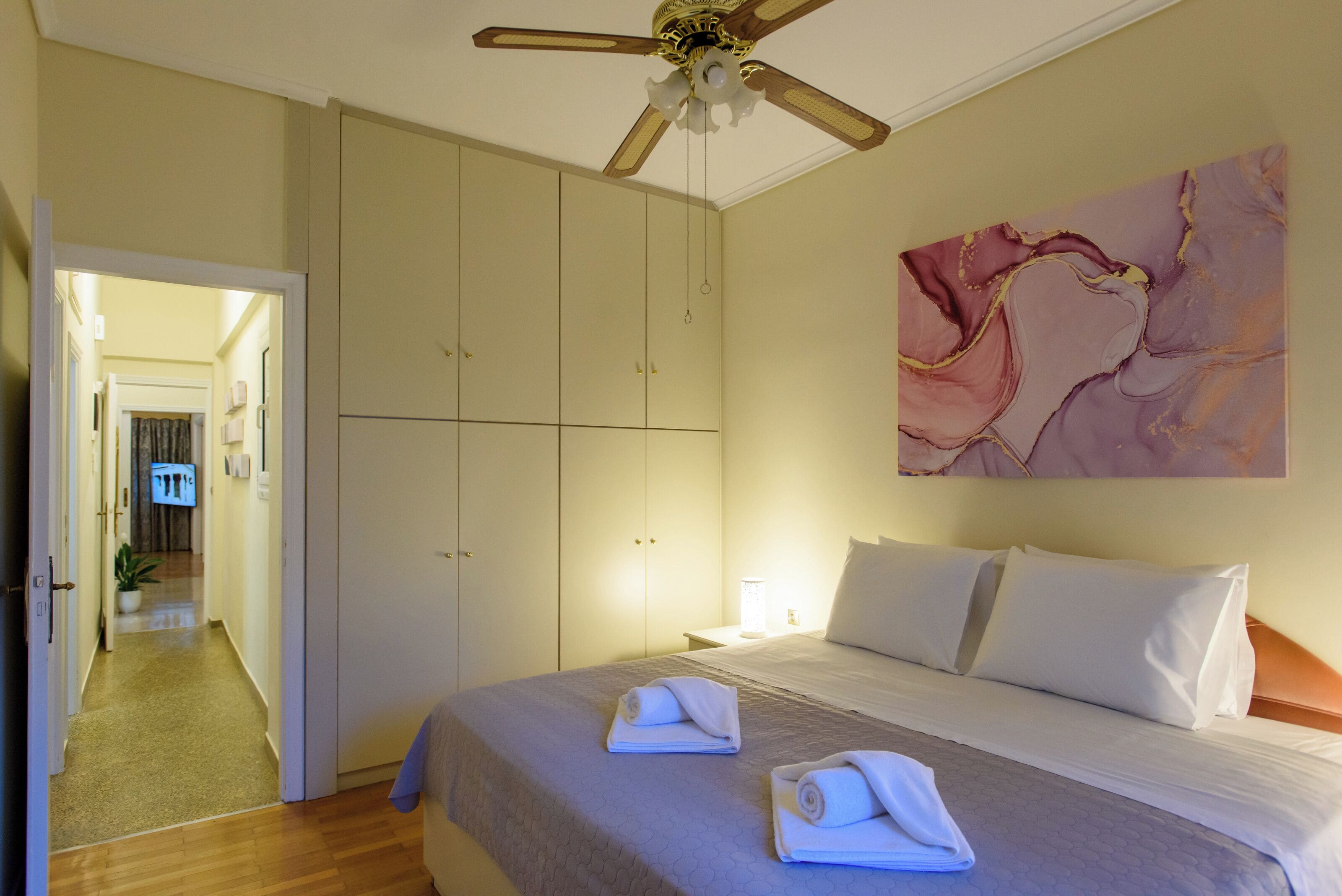 A hotel room with a bed, a ceiling fan, a painting on the wall, and a hallway leading to another room.