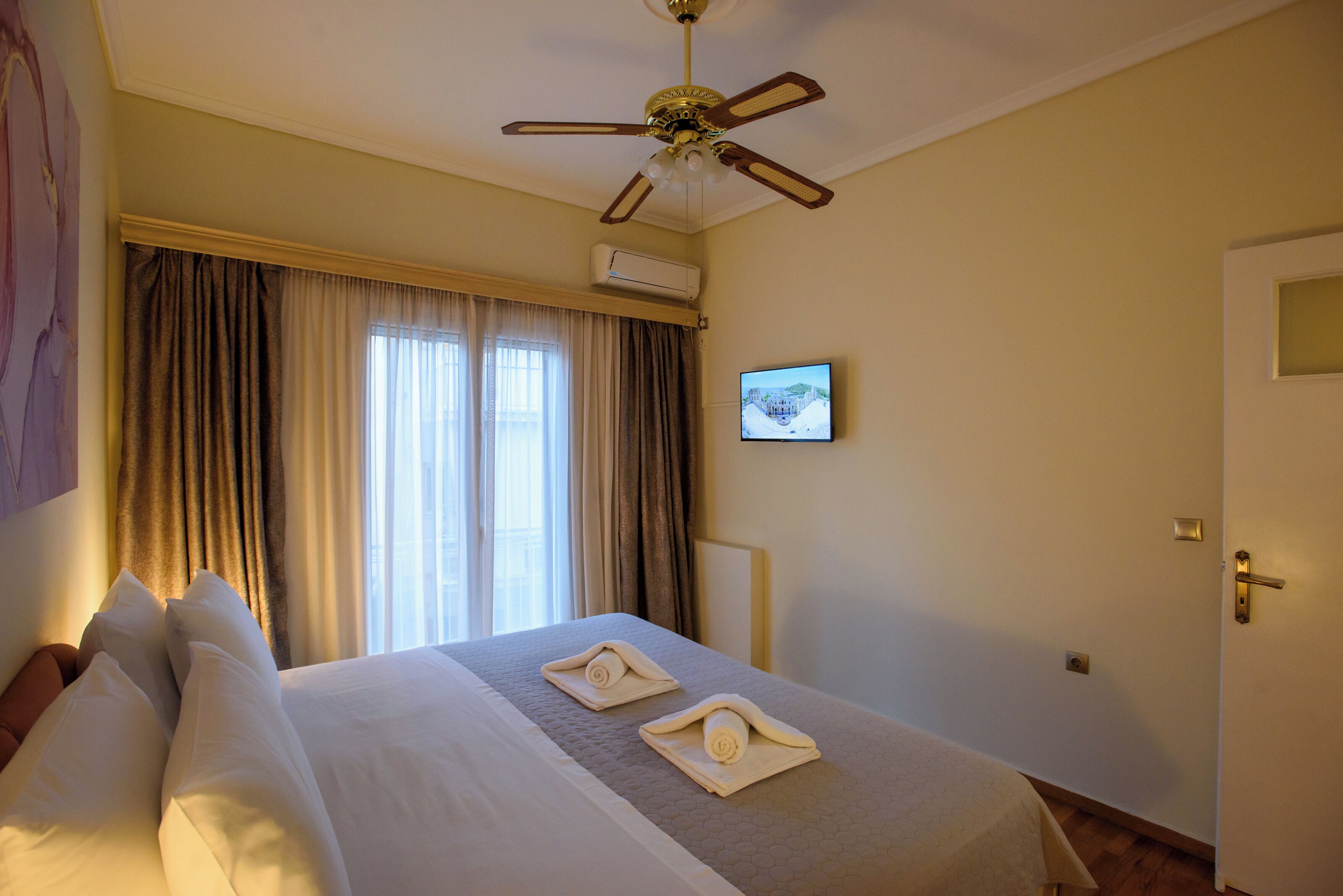 Habitación de hotel con cama, cortinas, ventilador de techo, aire acondicionado, televisor y puerta.