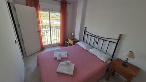A bedroom with a bed, a nightstand, a desk, a chair, and a view of the outside.