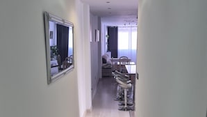 A narrow hallway with a light-colored wooden floor, a mirror on the left wall, and a dining area with a table and chairs in the distance.