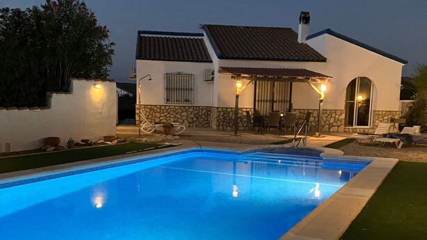 Una piscina con borde de piedra, una casa blanca con techo de tejas e iluminación exterior.