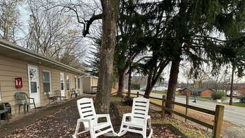 A row of white rocking chairs under a large tree, a building with beige siding, and a wooden fence.