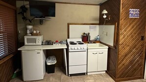 Cocina compacta con microondas, estufa y refrigerador, un pequeño bote de basura y una pared de madera con un tablón de anuncios.