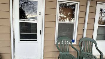 A house with a white door marked with the number 2, a small porch with two green plastic chairs, and a window with a view of trees.