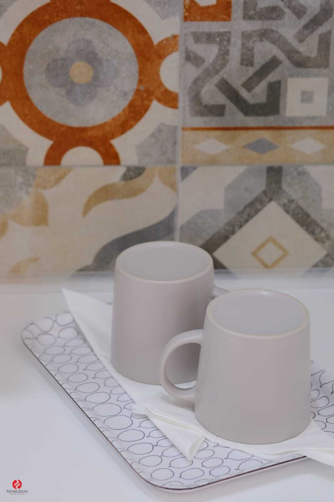 Deux tasses en céramique blanche, posées sur une serviette blanche ornée d’un motif circulaire, sont disposées sur une surface blanche dont le mur arrière est recouvert de carreaux aux motifs géométriques.