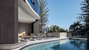 A modern pool area with lounge chairs and a clear pool, surrounded by buildings and trees. - Vue Broadbeach - Official (Broadbeach)