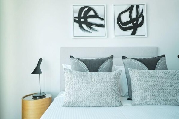 Une chambre à coucher bien rangée, avec un lit, des oreillers, une lampe de chevet et une œuvre d’art abstraite accrochée au mur.