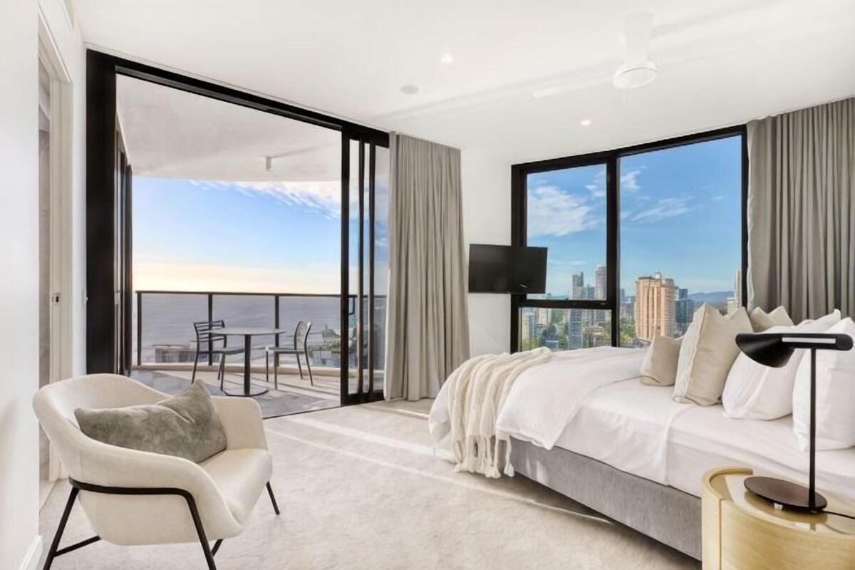 A modern hotel room with a large bed, a TV, a balcony with a city view, and a dining area.