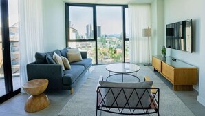 A modern living room with a sofa, coffee table, and TV, large windows with a city view, and a sliding door. - Vue Broadbeach - Official (Broadbeach)