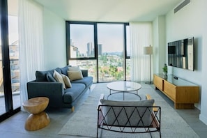 Una sala de estar moderna con sofá, mesa de centro y televisor, ventanas amplias con vista a la ciudad y un balcón.