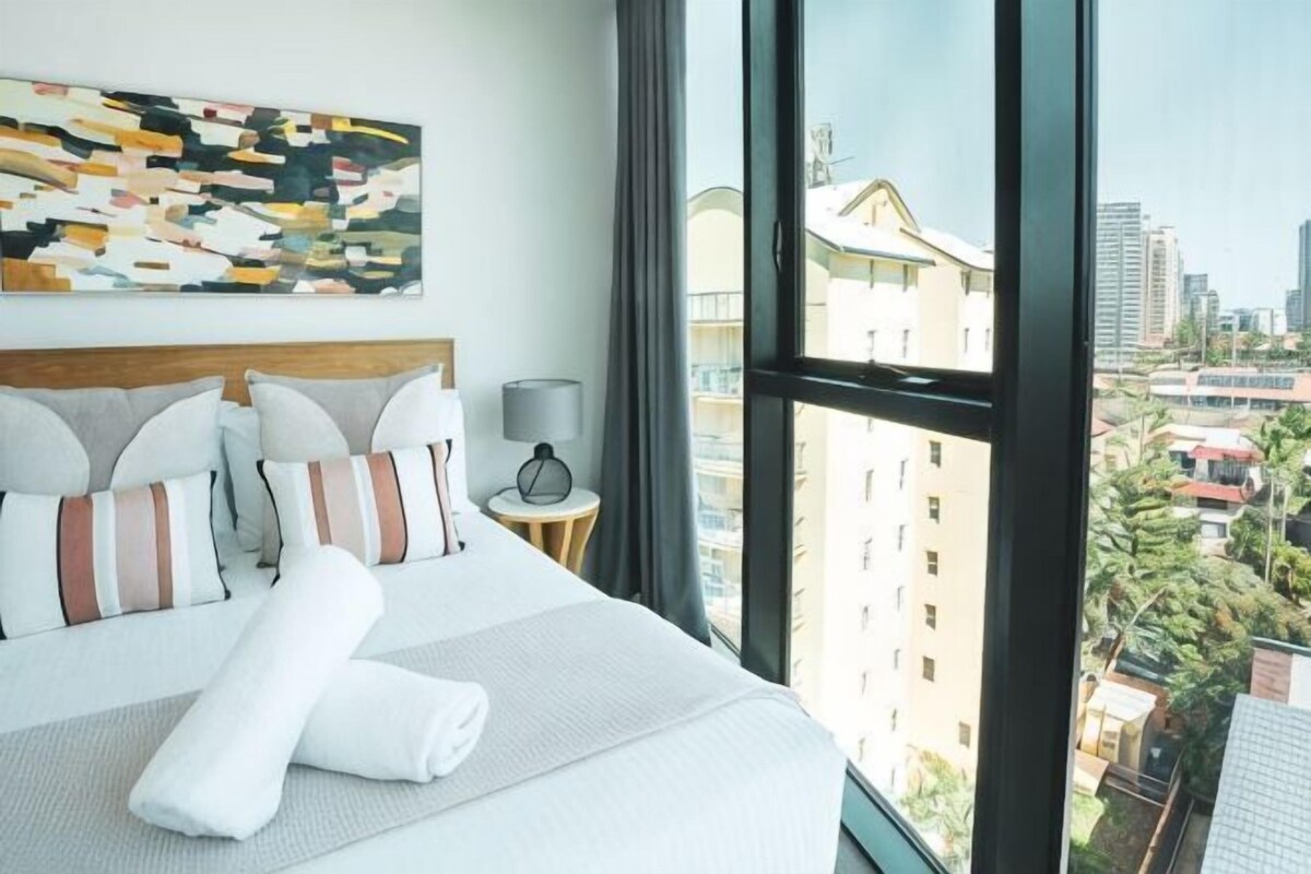 A hotel room with a large bed, a nightstand, a lamp, and a city view.