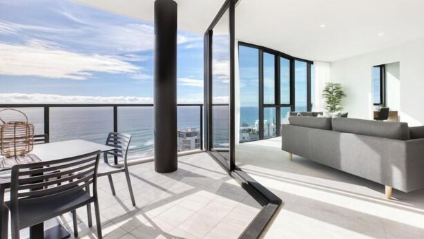 A modern living room with a large window, a sofa, and a dining area with chairs. - Vue Broadbeach - Official (Broadbeach)