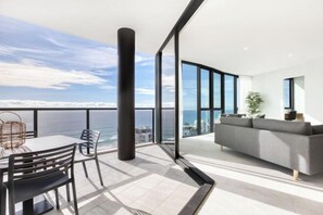 A modern living room with a large window, a sofa, and a dining area with chairs. - Vue Broadbeach - Official (Broadbeach)