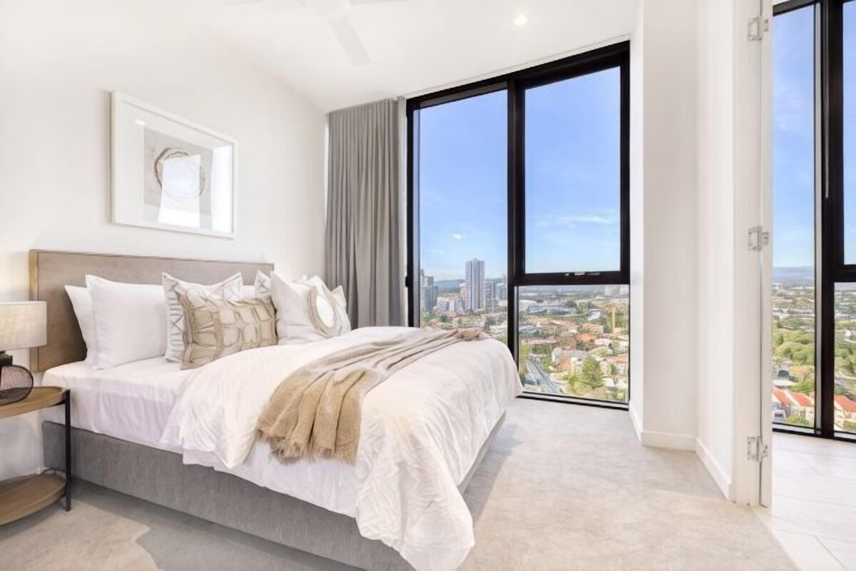 A bedroom with a large bed, a nightstand, a lamp, and a city view through large windows.
