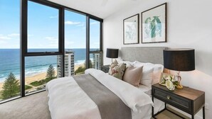 A bedroom with a large window overlooking the ocean, a bed, a nightstand with a lamp, and framed artwork on the wall. - Vue Broadbeach - Official (Broadbeach)