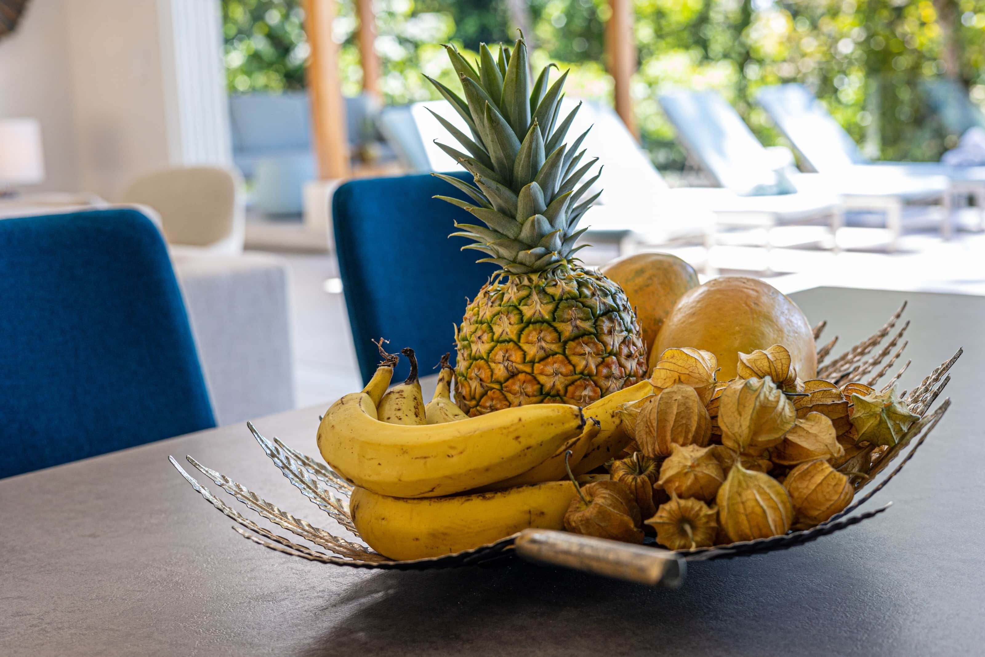 Una mesa con un frutero que contiene una piña, plátanos y flores secas.