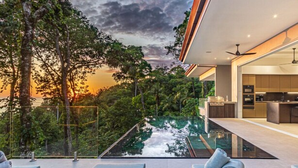 Ein moderner Außenbereich mit einem Pool mit Glasboden, Liegestühlen und Blick auf den Sonnenuntergang über einer üppigen Landschaft.