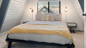 A bedroom with a sloped ceiling, a bed with a yellow and white quilt, two wall-mounted lamps, and a view of a house through a large window.