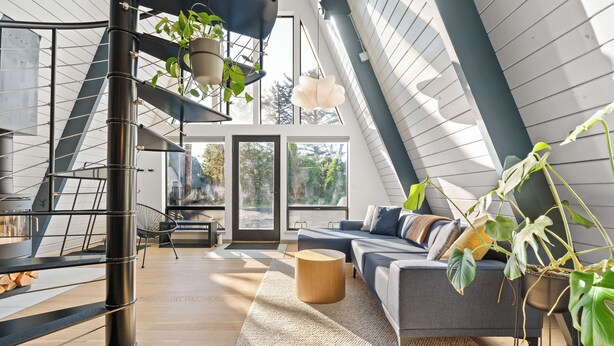 A modern loft with a high ceiling, a staircase with plants, a grey sofa, and a large window offering a view of trees.