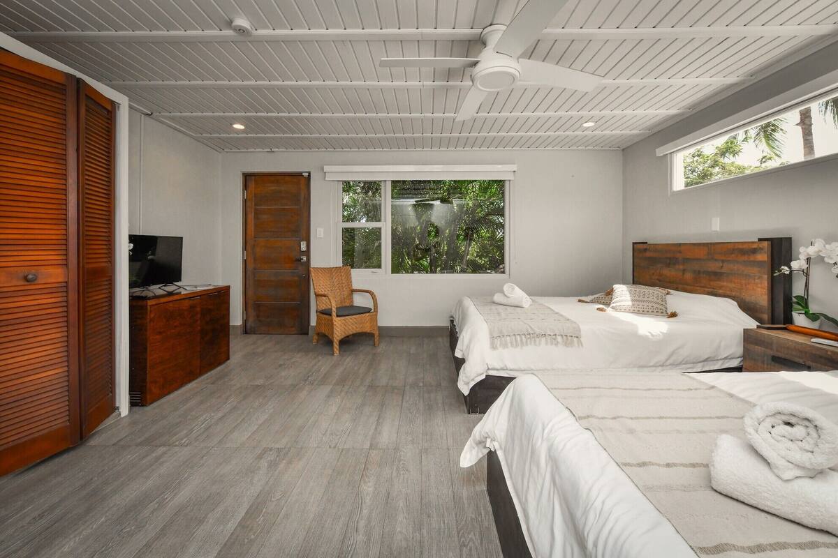 A modern bedroom with two beds, a wooden wardrobe, a television, and a large window with a view of greenery.