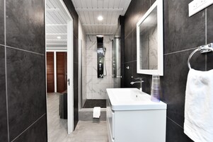 Un baño moderno con un lavamanos blanco, paredes de azulejos oscuros y un espejo grande.