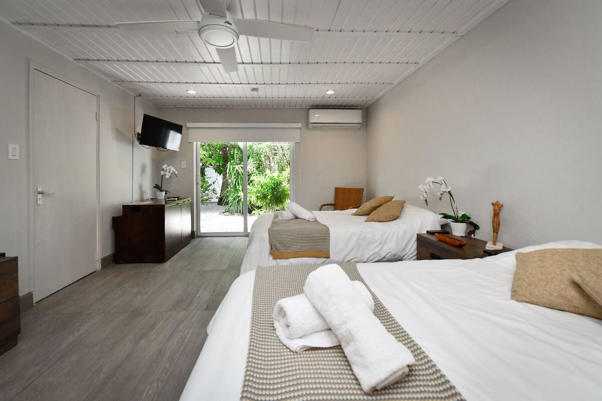 A modern hotel room with two beds, a TV, a desk, and a view of greenery.