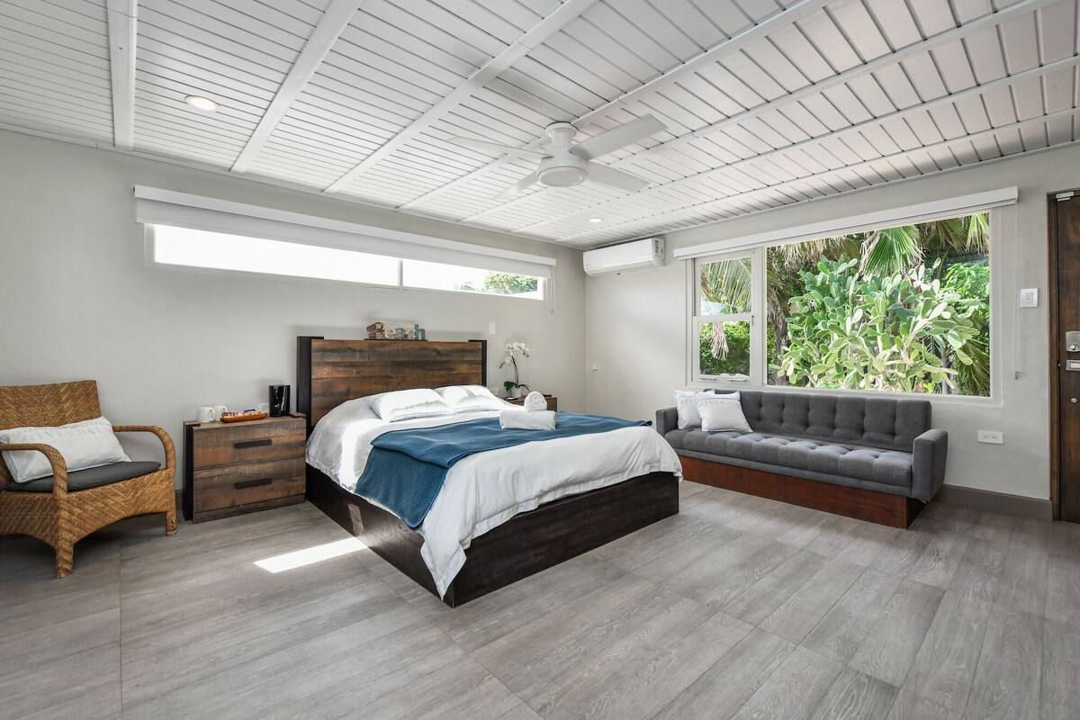 A modern bedroom with a large bed, a grey sofa, a wicker chair, and a window with a view of greenery.
