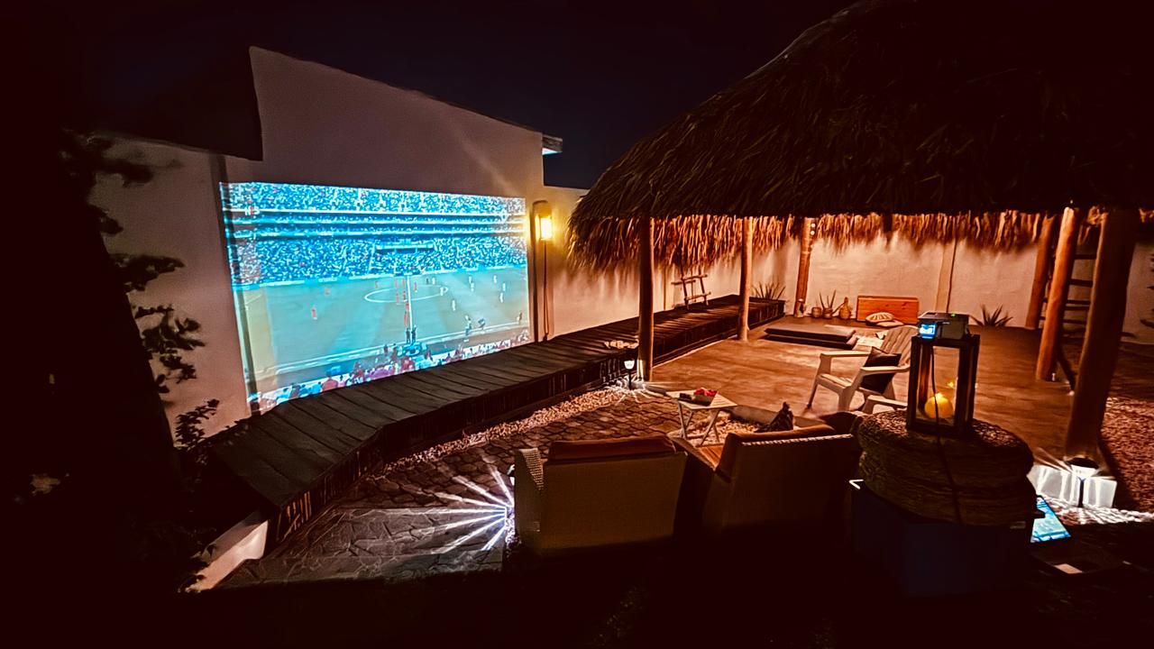 Un espace extérieur doté d’un toit de chaume, avec un écran de projection diffusant un événement sportif, des fauteuils confortables et une petite table avec une lampe.