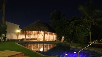 A pool area with a thatched-roof structure, lounge chairs, and palm trees.
