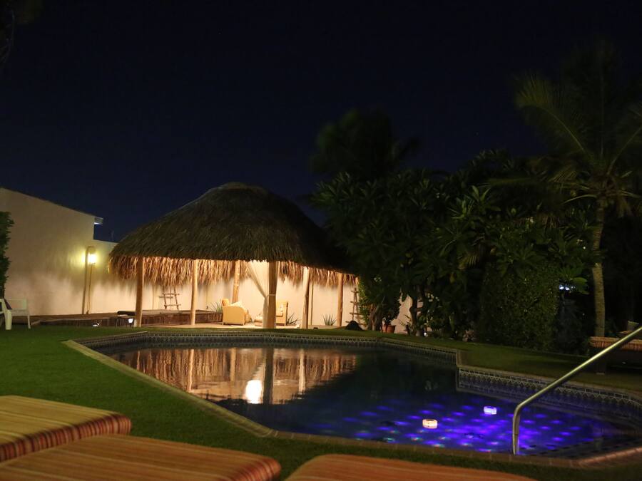 A pool area with a thatched-roof structure, lounge chairs, and palm trees.