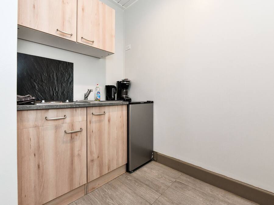 A compact kitchen with wooden cabinets, a stainless steel refrigerator, and a countertop with a coffee maker and a bottle of dish soap.