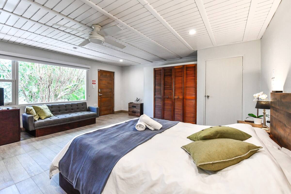 A modern bedroom with a large bed, a sofa, and a window with a view of trees.