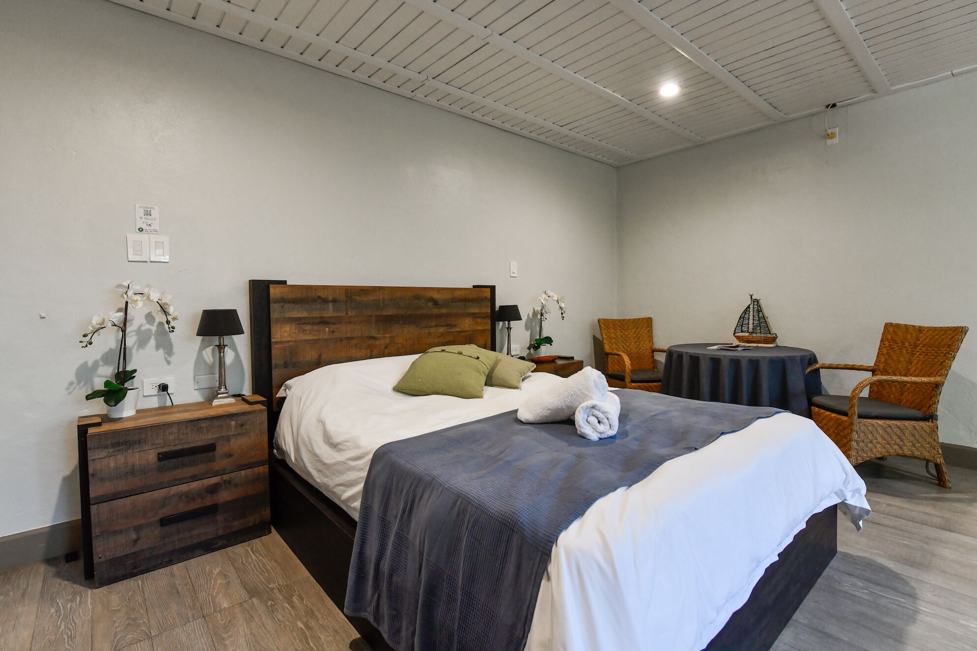 Ein Schlafzimmer mit einem Bett mit Holz-Kopfteil, einem Nachttisch mit Lampe, einem kleinen runden Tisch mit Stuhl und einer Orchideenpflanze.
